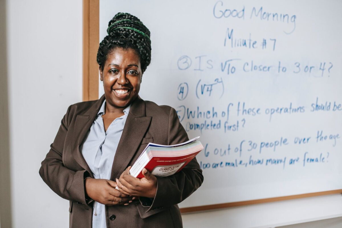 cheerful black female teacher with workbooks standing near whiteboard